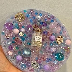 May include: A round, resin art piece with a mix of colorful beads, gems, and charms. A small glass bottle with a cork stopper and the text "Rose Quartz Quartz Rose" is in the center. Gold-colored charms include a tree of life, a lotus flower, and a hamsa hand.