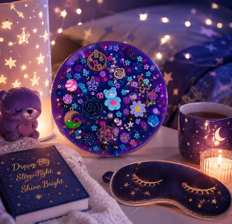 May include: A circular purple resin art piece embedded with various charms including a blue bear, flowers, stars, and a crescent moon. The piece is surrounded by a purple book with gold text, a purple sleep mask, a mug, and a star-patterned lamp.