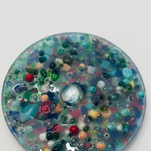 Peut inclure: Un dessous de verre rond en r&eacute;sine, orn&eacute; d'un m&eacute;lange de perles et d'&eacute;l&eacute;ments d&eacute;coratifs color&eacute;s. Le dessous de verre pr&eacute;sente des couleurs vari&eacute;es, notamment le bleu, le vert, le rouge et le blanc, avec une grosse perle argent&eacute;e au centre.