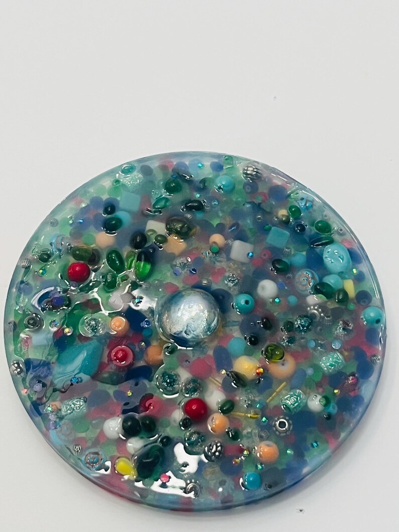 Peut inclure: Un dessous de verre rond en r&eacute;sine, orn&eacute; d'un m&eacute;lange de perles et d'&eacute;l&eacute;ments d&eacute;coratifs color&eacute;s. Le dessous de verre pr&eacute;sente des couleurs vari&eacute;es, notamment le bleu, le vert, le rouge et le blanc, avec une grosse perle argent&eacute;e au centre.