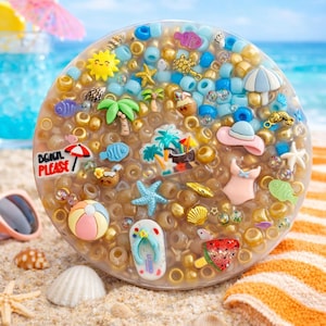 May include: A clear, round resin piece filled with gold and blue beads, and miniature beach-themed charms. Charms include a sun, palm trees, starfish, and a sign that says "Beach Please". The piece is on a sandy surface.