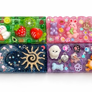 May include: Four rectangular resin trays featuring embedded decorative elements. The top left is green with fruit charms. The top right is pink with a rabbit and bear. The bottom left is dark blue with a sun and moon. The bottom right is purple with animal figures.