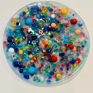 Peut inclure: Une &oelig;uvre d'art en r&eacute;sine ronde et transparente, remplie de perles et d'ornements color&eacute;s. La pi&egrave;ce pr&eacute;sente une vari&eacute;t&eacute; de couleurs, dont le bleu, le rouge, le jaune et l'orange. Les perles sont de tailles et de formes vari&eacute;es, cr&eacute;ant un design textur&eacute; et vibrant.