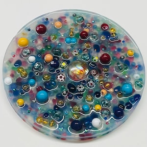 Peut inclure: Un dessous de verre rond en r&eacute;sine transparente, incrust&eacute; de perles color&eacute;es et d'&eacute;l&eacute;ments d&eacute;coratifs. Le dessous de verre pr&eacute;sente un m&eacute;lange de perles bleues, rouges, jaunes et transparentes, avec un marbre de verre iris&eacute; au centre.