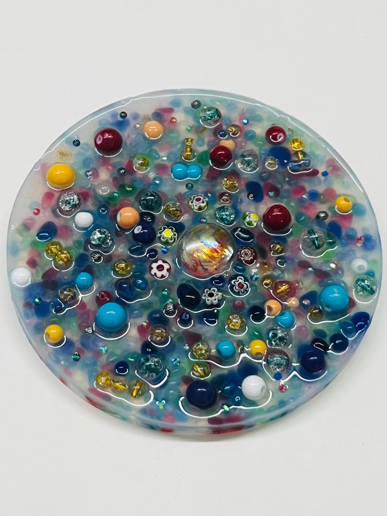 Peut inclure: Un dessous de verre rond en r&eacute;sine transparente, incrust&eacute; de perles color&eacute;es et d'&eacute;l&eacute;ments d&eacute;coratifs. Le dessous de verre pr&eacute;sente un m&eacute;lange de perles bleues, rouges, jaunes et transparentes, avec un marbre de verre iris&eacute; au centre.
