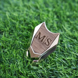以下が含まれることがあります： 木製の銀色のリングで、木の部分に「M|S」のイニシャルが刻まれています。リングは緑の芝生の上に置かれています。
