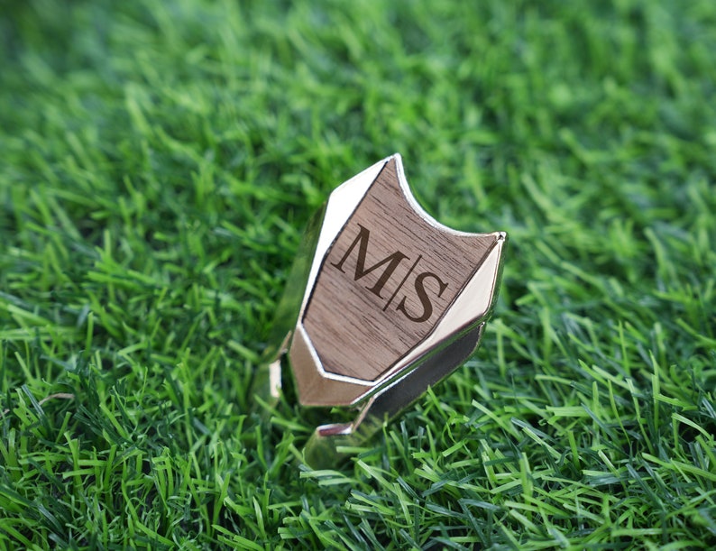 以下が含まれることがあります： 木製の銀色のリングで、木の部分に「M|S」のイニシャルが刻まれています。リングは緑の芝生の上に置かれています。