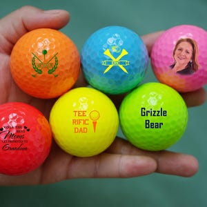 May include: Six golf balls with different designs. The golf balls are orange, blue, pink, red, yellow, and green. The designs include text, a golf club, and a picture of a woman.