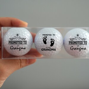 May include: Three white golf balls in a clear plastic box. Each ball has black text that reads "Promoted to Grandma" with a design of a flower or footprints.
