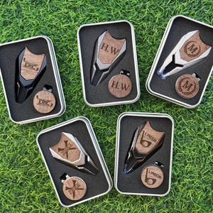 Op de afbeelding: Vijf zwarte en zilveren golfbalmarkers met gepersonaliseerde houten inlegstukken. Elke marker bevindt zich in een aparte zwarte doos met een zilveren rand. De dozen bevinden zich op een groene grasachtergrond.