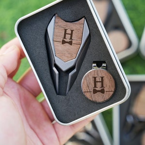 以下が含まれることがあります： 金属製のクリップと、パーソナライズされたイニシャル「H」と蝶ネクタイのデザインが施された木製ゴルフボールマーカー。マーカーは黒いベルベットのギフトボックスに入っています。