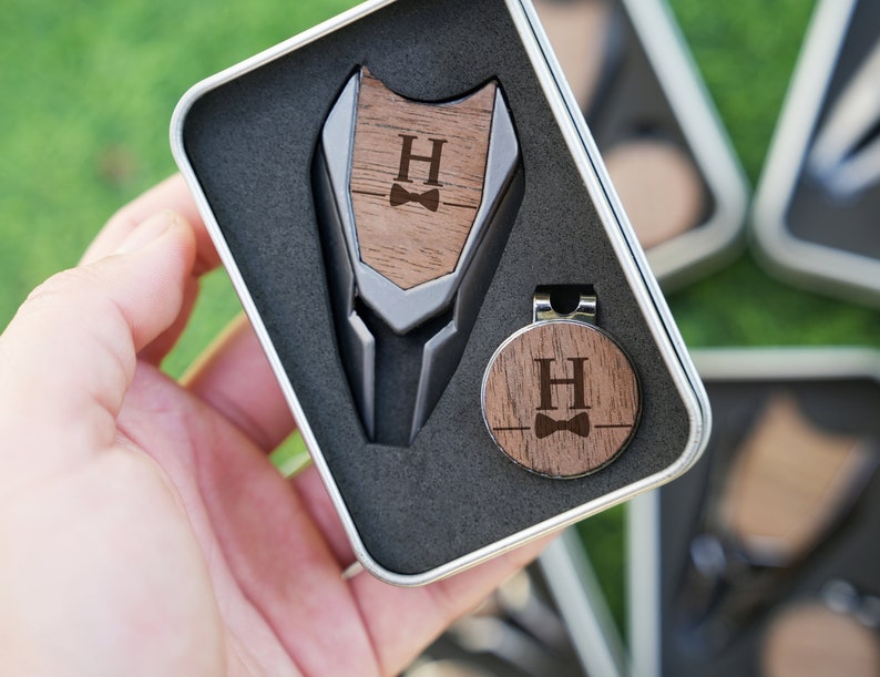 以下が含まれることがあります： 金属製のクリップと、パーソナライズされたイニシャル「H」と蝶ネクタイのデザインが施された木製ゴルフボールマーカー。マーカーは黒いベルベットのギフトボックスに入っています。