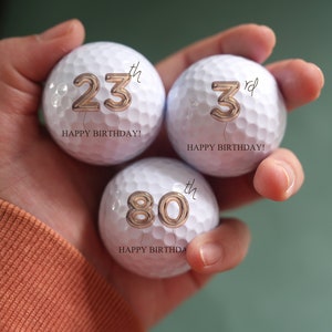 Könnte beinhalten: Drei weiße Golfbälle mit goldenen Zahlen und dem Schriftzug "Happy Birthday!" darauf. Die Zahlen sind 23., 3. und 80.