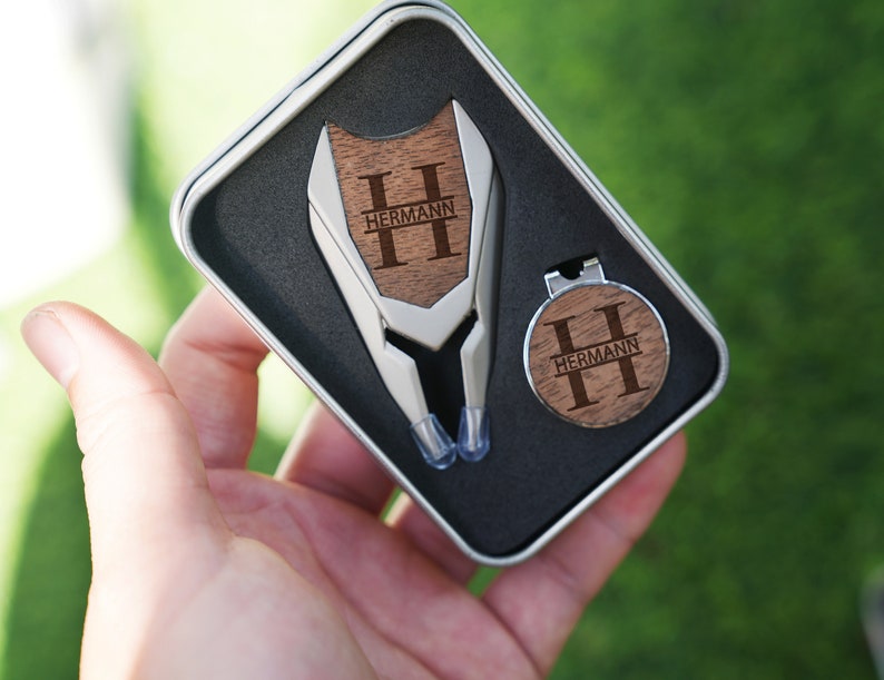以下が含まれることがあります： 黒いギフトボックスに入った、木製のハンドルに「HERMANN」と刻まれた銀製のゴルフツールセットです。