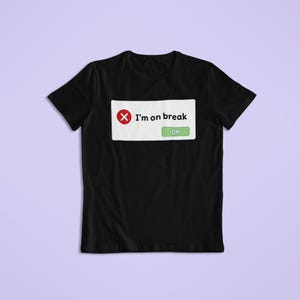 May include: Black t-shirt with a white box displaying the text "I'm on break" and an "OK" button. A red "X" symbol is also present. The shirt is set against a light purple backdrop.