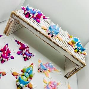 May include: A collection of colorful, handmade polymer clay dragon figurines. The dragons feature a variety of vibrant colors, including pink, blue, yellow, and green. Some dragons are arranged on a small, white-washed wooden shelf.