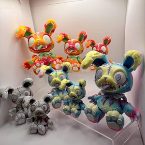May include: A collection of seven rabbit figurines in two distinct color schemes. Four figures feature vibrant orange, yellow, and green tones, while three figures display pastel blue, pink, and yellow hues. Each rabbit has stitched details, spiral eyes, and an open mouth with a tongue.