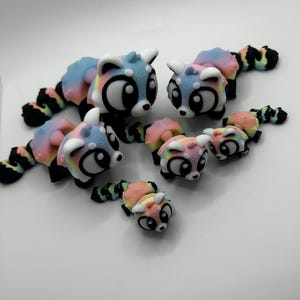 Peut inclure: Plusieurs figurines de pandas roux stylisés et colorés. Chaque figurine présente un dégradé de couleurs pastel sur le corps et la queue, avec des accents noirs et blancs. Les figurines varient en taille, avec des versions grandes et petites.