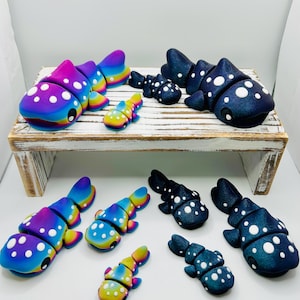 Può includere: Una collezione di sculture di pesci colorate e stilizzate. I pesci hanno un design del corpo segmentato con pois bianchi. La tavolozza dei colori comprende tonalità vibranti di blu, viola, giallo e verde. Alcuni sono esposti su una mensola di legno rustica.