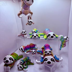 May include: A collection of colorful sloth figurines in various poses. The sloths are in shades of rainbow, with white faces, black eyes, and white claws. Some sloths are hanging, while others are lying down or stacked. The figurines are displayed on clear acrylic shelves.