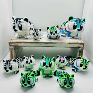May include: A selection of cartoon leopard figurines in various colours. The figurines have large eyes and come in white with black spots, green, and rainbow shades. Some are displayed on a small wooden shelf.