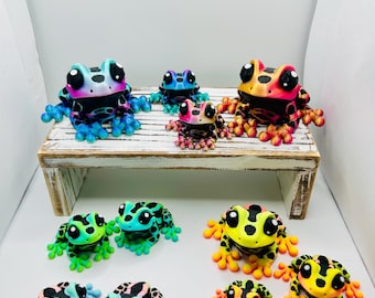 Peut inclure: Une collection de onze figurines de grenouilles articulées de petite taille, aux dégradés de couleurs variés comme le bleu, le rose, l'orange, le jaune et le vert, avec des taches noires et de grands yeux sombres, disposées sur un banc en bois blanc.
