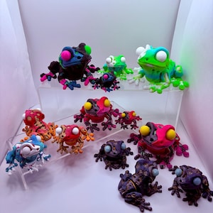 May include: A collection of articulated frog figurines in various sizes and color combinations, including black with neon pink, green with blue, red with yellow, and purple with gold. Each frog features large, round eyes and jointed limbs.