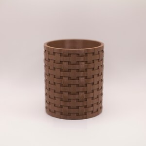 May include: A brown cylindrical container with a woven basket-like texture. The container is made of a solid material and has a smooth interior. The design gives the appearance of a basket. The container is suitable for holding pencils, pens, or small items.