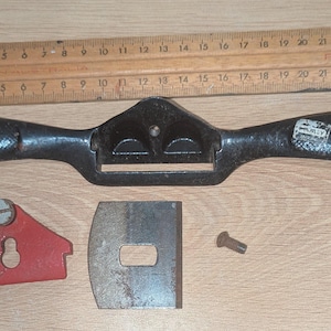 May include: A black metal spokeshave tool with a textured handle, positioned below a wooden ruler measuring 26 centimetres. Included are a red metal cap with a screw, a rectangular steel blade, and a small metal bolt, all laid out separately.