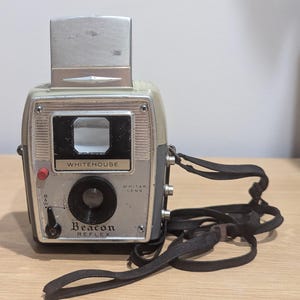 May include: A vintage Whitehouse Beacon Reflex camera featuring a beige and silver body with a black lens, a top-mounted viewfinder, and a black fabric neck strap. The front panel displays the text Whitehouse, Beacon Reflex, and Whitar Lens.