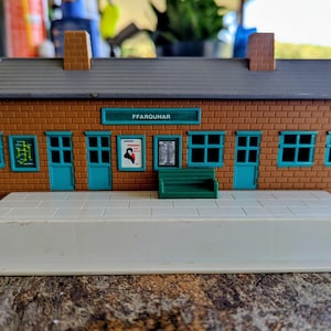 May include: A model train station building with brown brick walls, a grey roof, and two chimneys. The structure features teal doors, window frames, and a sign reading FFARQUHAR. A white platform extends from the front, and a small green bench is attached.