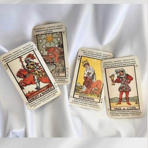 May include: Four tarot cards arranged on a white fabric surface. The cards are Knight of Wands, The Star, Strength, and Page of Cups. Each card features a colorful illustration and printed text describing themes like adventure, inspiration, loyalty, and innocence.