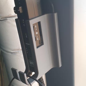 May include: A silver-colored device with a black component, featuring buttons, ports, and the text "PRIORIT" and "UFC". The device has a hook for holding a headset or similar item. The device is mounted on a wall.