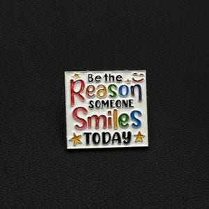 Peut inclure: Épinglette carrée en émail avec les mots "Be the Reason Someone Smiles Today" en lettrage coloré. L'épinglette a un fond blanc avec une bordure argentée et des étoiles.