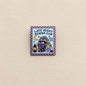 May include: A rectangular enamel pin shaped like a postage stamp with a light blue background. It features a raccoon reading a book while wrapped in a colourful patchwork quilt, a yellow crescent moon, a lantern, and the text LATE NIGHT READER CLUB.
