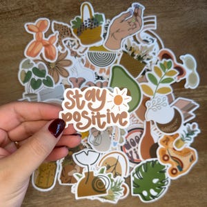 May include: A collection of stickers featuring various designs, including a hand holding flowers, a potted plant, a sun, an avocado, a roller skate, and a sticker that says "Stay Positive".