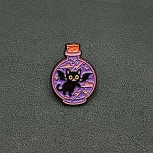 May include: A purple potion bottle pin featuring a black cat with bat wings. The pin has a pink border, a cork top, and lightning bolt and star accents. The cat has large eyes and is smiling.