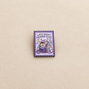 Peut inclure: Une broche en émail rectangulaire en forme de timbre-poste avec un bord festonné. Le motif représente un hibou brun portant des lunettes, lisant un livre sous une couette étoilée. Le texte indique LATE NIGHT READER CLUB et SLEEP IS OPTIONAL BOOKS BETTER.