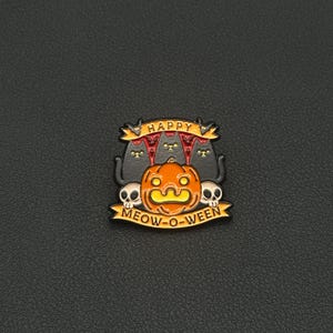 May include: A Halloween enamel pin with a pumpkin, black cats, and skulls. The pin reads "HAPPY" and "MEOW-O-WEEN" on banners. The design is in orange, black, white, and yellow.