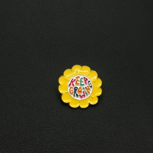 Peut inclure: Épingle en émail jaune en forme de fleur avec les mots "Keep Growing" en lettres colorées. L'épingle a un centre blanc et un bord festonné.