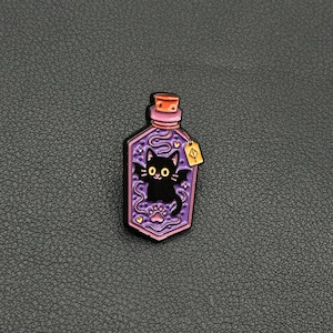 Peut inclure: Épingle en émail violet en forme de flacon de potion, représentant un chat noir aux yeux jaunes et une empreinte de patte. La bouteille a un bouchon en liège et une petite étiquette. L'épingle est posée sur un fond sombre.