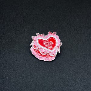 May include: A pink and red heart-shaped enamel pin with the text "literally just a girl". The pin features a layered design with white trim and pink bows.