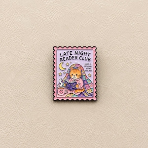 Peut inclure: Une broche en émail rectangulaire en forme de timbre-poste avec un fond violet clair. Elle représente un chat orange lisant un livre sous une courtepointe colorée, une tasse fumante, un croissant de lune et des étoiles. Texte : LATE NIGHT READER CLUB. SLEEP IS OPTIONAL. BOOKS ARE NOT.