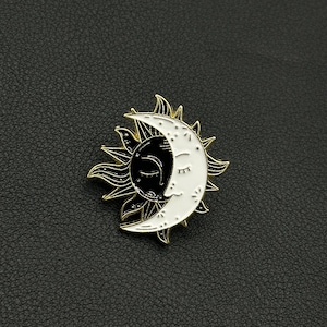 Peut inclure: Épingle à nourrice émaillée sur le thème céleste, représentant un soleil et une lune. Le soleil est noir avec des rayons dorés, et la lune est blanche avec un visage endormi. La broche mesure environ 2,5 cm de diamètre.