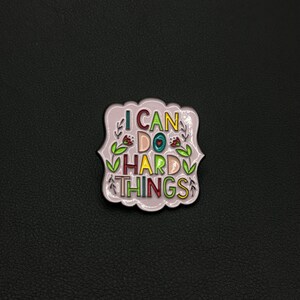 Peut inclure: Épingle en émail violet clair avec les mots "I CAN DO HARD THINGS" en lettres colorées. L'épingle a une bordure décorative et des motifs floraux. L'épingle est sur une surface noire.
