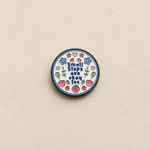 May include: A round enamel pin with a white background and a dark blue border. The centre features the blue text Small Steps are okay too, surrounded by colourful illustrations of pink hearts, blue flowers, orange stars, green leaves, and a strawberry.