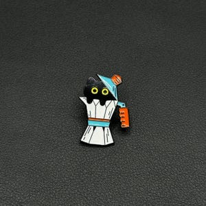 May include: A black cat pin with yellow eyes and a blue hat sits on a white and blue espresso maker. The pin has an orange handle. The pin is set against a black background.