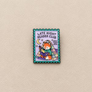 Peut inclure: Une broche émaillée rectangulaire en forme de timbre-poste avec une bordure sarcelle. Elle représente un renard orange lisant un livre sous une couette colorée avec une lampe de poche. Le texte indique LATE NIGHT READER CLUB et SLEEP IS OPTIONAL BOOKS ARE NOT.