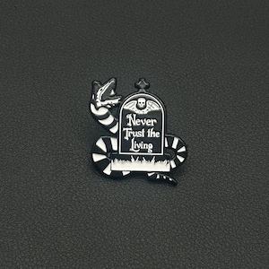 May include: A black and white enamel pin depicting a snake wrapped around a tombstone. The tombstone features a skull and the words "Never Trust the Living".
