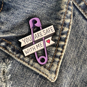 May include: Purple safety pin enamel pin with a white banner that reads "You are safe with me" and a small red heart.